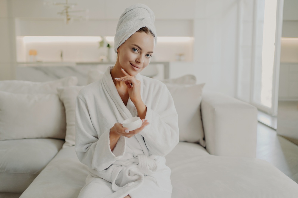 Portrait of young pretty woman with in white bathrobe and towel on head holding cosmetic cream, touching her soft healthy face skin and smiling at camera while relaxing on sofa in cozy apartment. Young beautiful woman holding cosmetic product and smiling while relaxing on sofa after taking shower at home