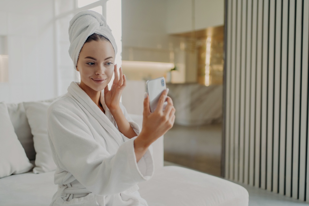 Beauty routine. Young gorgeous woman touching her soft healthy skin, applying cosmetic cream while sitting on sofa, female wearing bathrobe and towel on head holding smartphone in front of her face. Young beautiful woman applying cosmetic cream while relaxing on sofa after taking shower at home