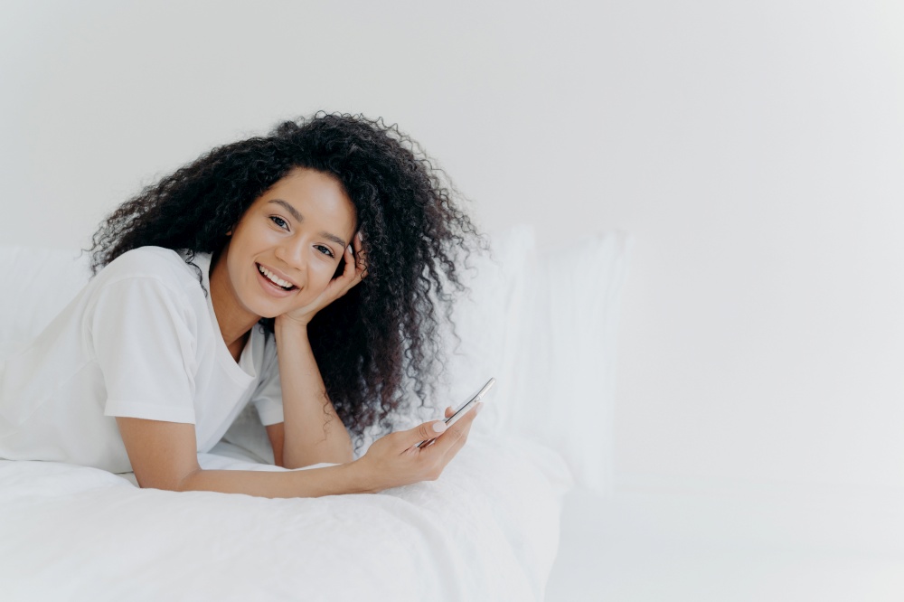 Overjoyed relaxed curly young African American woman enjoys spare time at home, lies in bed with modern cellphone, uses phone in bedroom, sends text message to lover, has lazy morning, good time