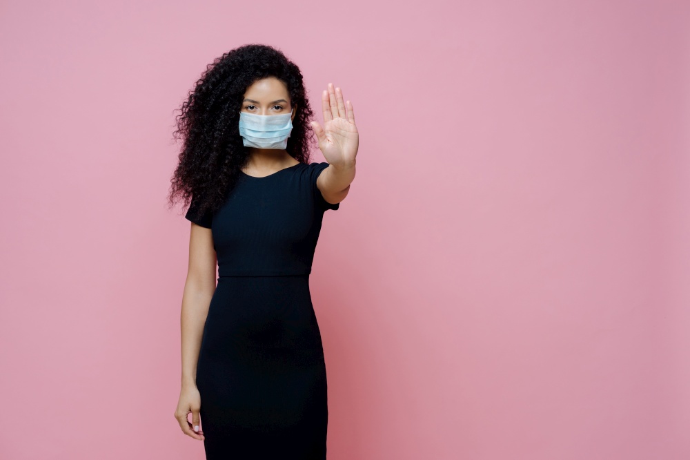 Covid 19 outbreak, viral disease. Photo of ethnic woman with curly hair makes stop gesture with palm, says no to coronavirus, wears protective sterile mask to avoid virus, dressed in black dress