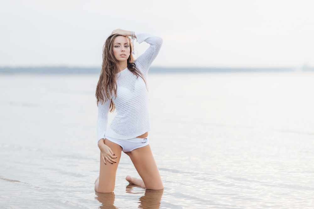Outdooor view of good looking brunette female with make up, dressed in white bikini, poses at camera near calm sea background, has fit body shape, feels free and relaxed. Woman at seashore