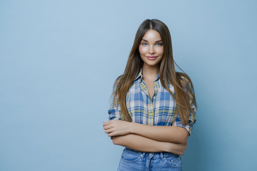Horizontal shot of attractive young female with long straight hair, wears checkered shirt, jeans, keeps hands crossed, has clean skin, minimal makeup, expresses positive emotions, enjoys photo session