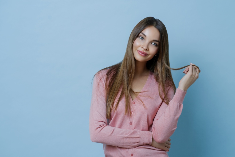 Waist up shot of pleasant looking young woman has beautiful long hair, tilts head, dressed in casual jumper, has perfect clean skin, advertises shampoo for hair care, isolated on blue background.