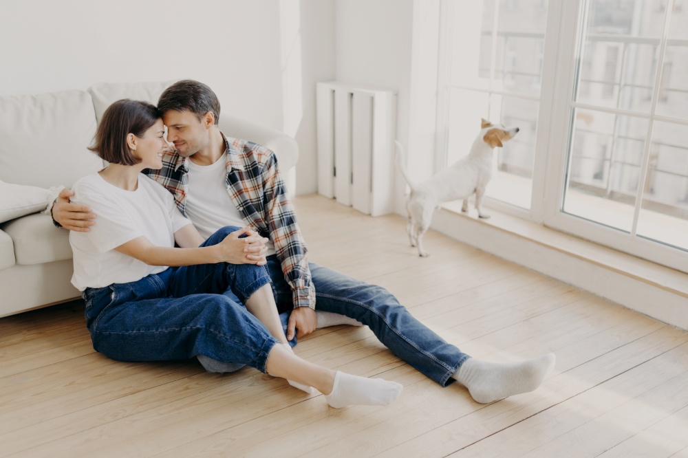 Happy newlywed couple move in new apartment, rejoice having spare time together, embrace and touch noses, going to have sweet kiss, pose on floor in empty room near sofa, their dog in background
