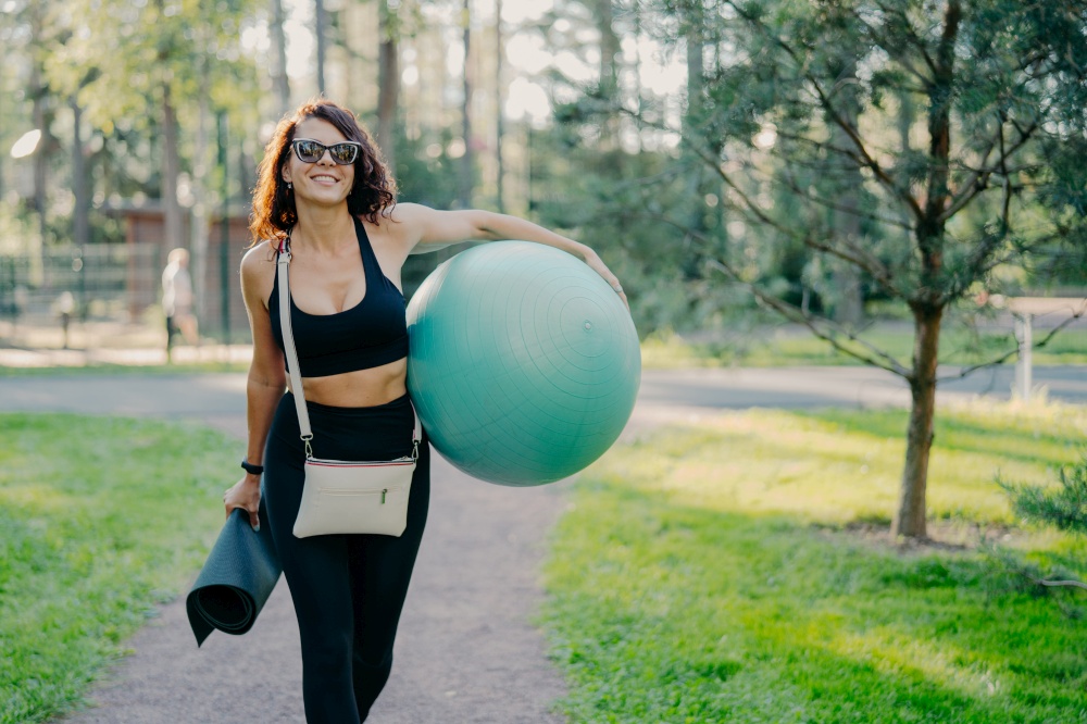 Outdoor shot of happy brunette slim woman dressed in active wear trendy sunglasses carries sport equipment walk on way enjoys fresh air and beautiful nature. Sporty female holds fit ball and karemat