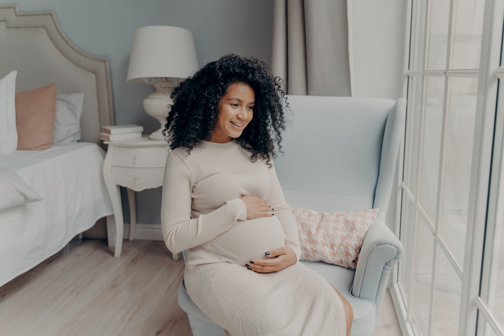 Charming young mixed race pregnant woman, future mother looking out of big window and dreaming about maternity while sitting sideways on armchair in bedroom, holding belly with both hands and smiling. Charming afro american pregnant woman looking in window and thinking about future baby