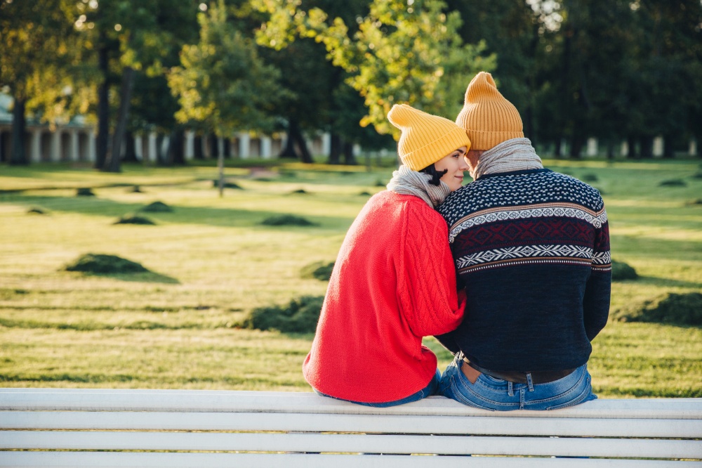Lovely couple sit on bench together, wear warm clothes and knitted hats, embrace each other, express love and good relationship, enjoy beautiful landscapes and fresh air in park. Relationship concept
