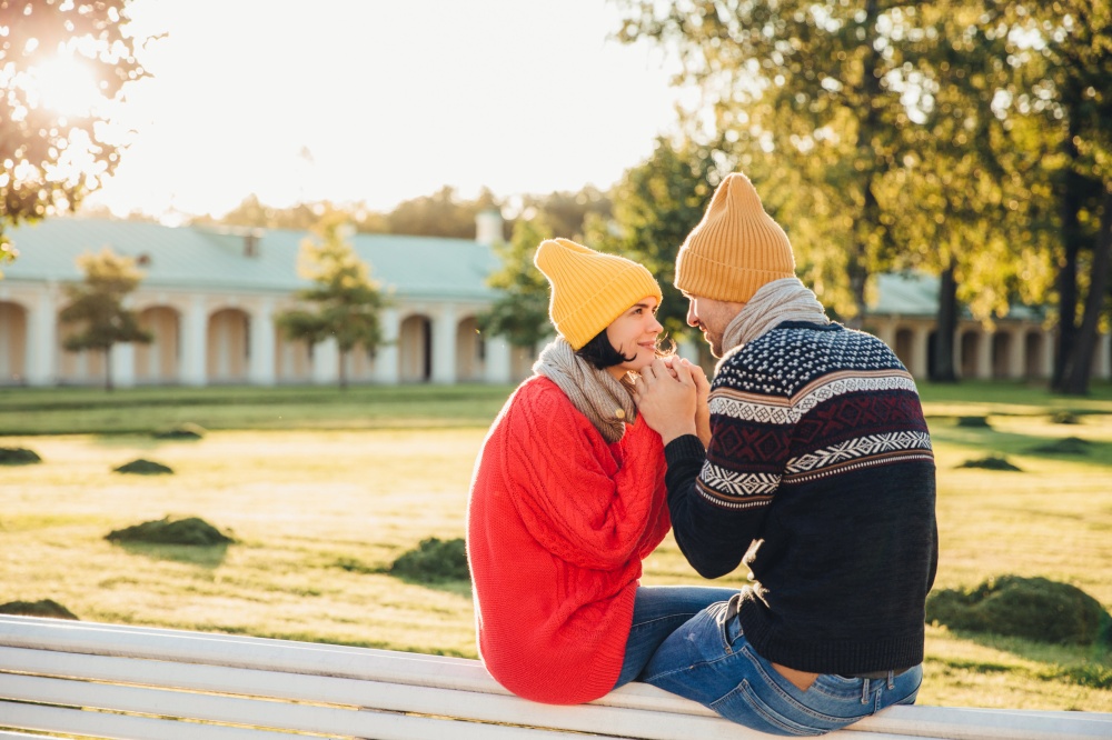 Adorable couple spend time together: attractive man hold his girlfriend`s hands, going to make her proposal, look with great love at her, sit on bench in park, feel relaxed, wear warm clothes