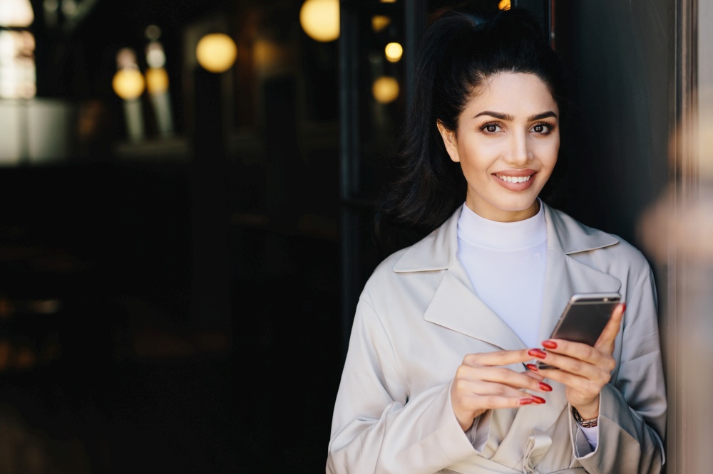 Attractive brunette businesswoman having dark hair tied in pony tail and wonderful make-up smiling demonstrating her white perfect teeth waitng for her business partner using modern smartphone