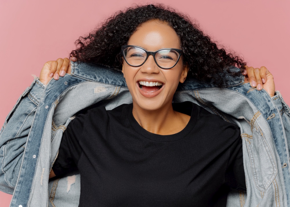 Isolated shot of curly cheerful woman tries on new denim jacket, wears optical glasses, smiles broadly, has perfect mood, poses against pink wall. Positive emotions. Afro female in stylish clothes