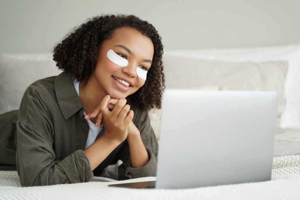 Face of carefree african american girl which applies eye patches and relaxing at home watching video on laptop. Attractive curly girl is grooming herself in bedroom and having fun. Beauty routine.. Face of carefree african american girl which applies eye patches and watching video on laptop.