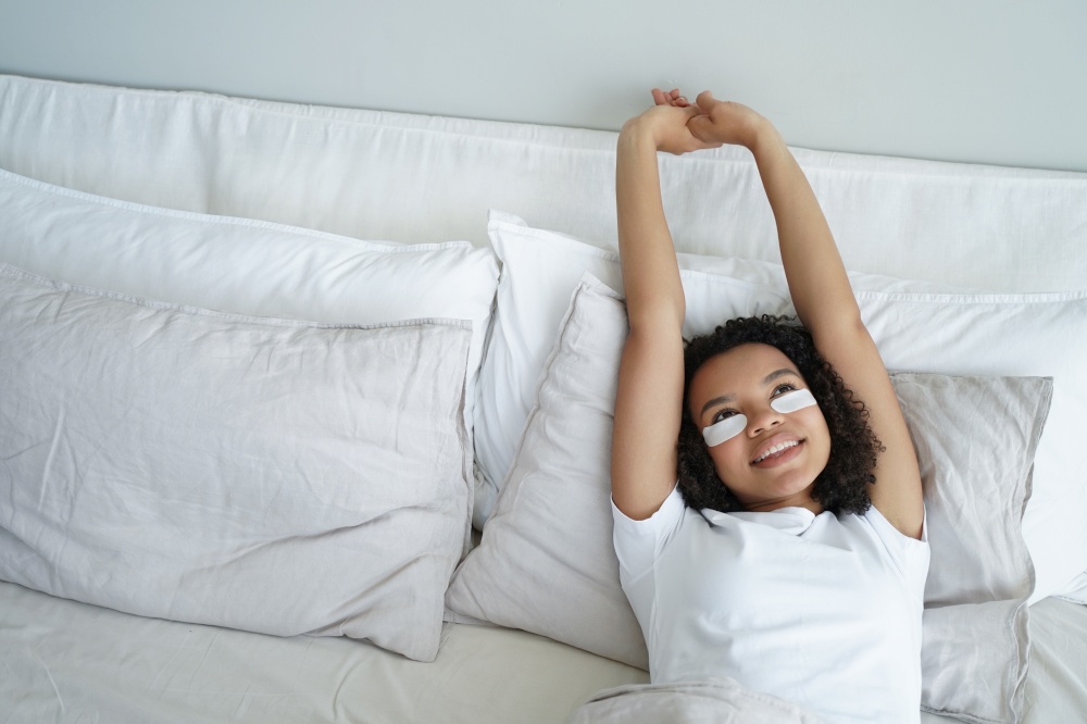 Young woman is stretching and relaxing in bed. Happy afro girl applies eye patches and relaxing at home enjoying weekend morning. Carefree spanish girl in bedroom. Comfort and leisure concept.. Happy young woman is stretching and relaxing in bed in morning. Comfort and leisure concept.