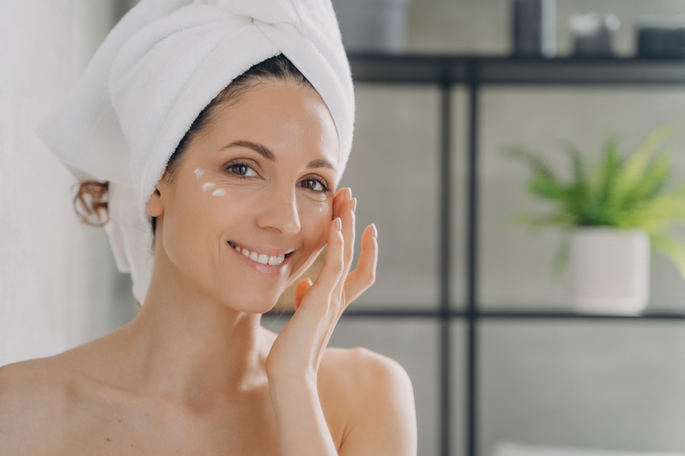 Face of beautiful european lady which applies cream. Attractive feminine caucasian girl wrapped in towel after bathing and hair washing. Happy young woman takes shower at home. Beauty routine.. Face of beautiful european lady which applies cream. Attractive feminine girl beauty routine.