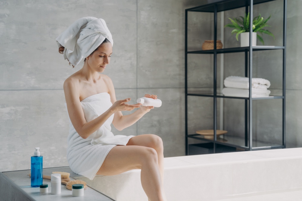 Beautiful european woman wrapped in towel applying body moisturizing cream after bathing. Young woman takes shower in morning at home. Anti-cellulite cosmetic, massage and bodycare.. Beautiful european woman wrapped in towel applying body moisturizing cream after bathing.
