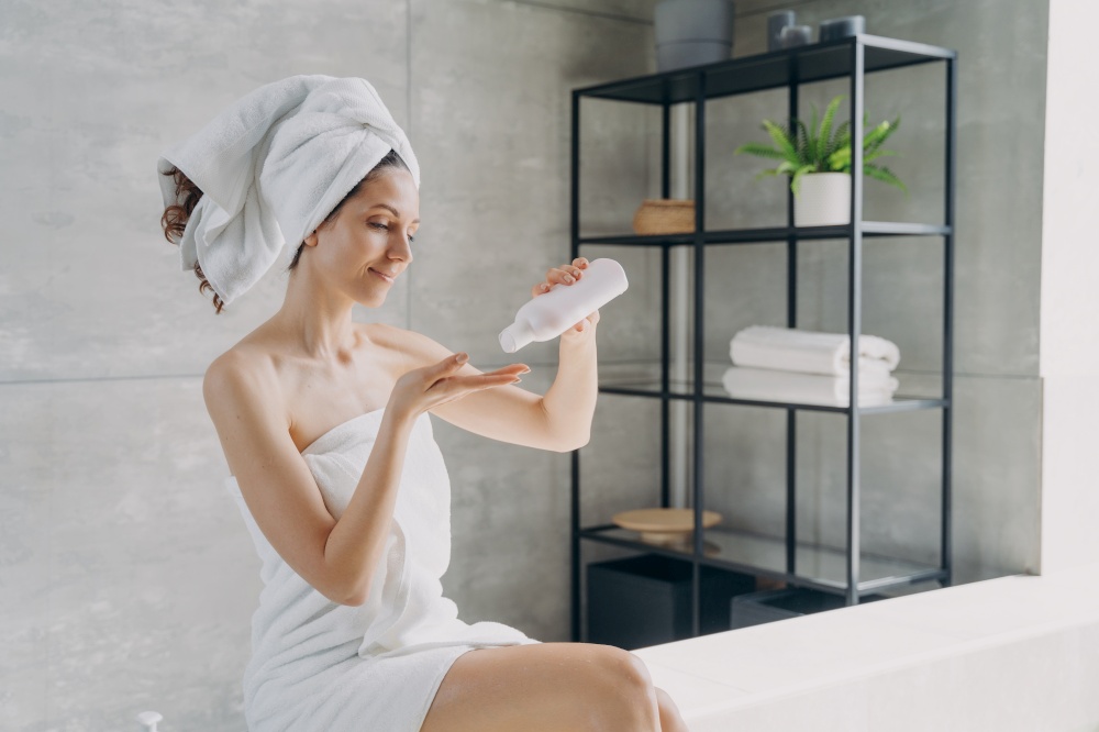 Body lotion applying from bottle. Attractive caucasian girl wrapped in towel after bathing. Happy young woman takes shower at home. Products for smooth silky skin using. Spa procedure.. Body lotion applying from bottle. Happy young woman using products for smooth silky skin.