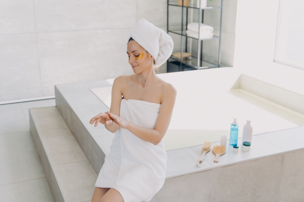 Girl applies eye patches and hand cream. Attractive caucasian woman wrapped in towel after bathing. Young hispanic lady takes shower at home. Relaxation at spa resort. Modern interior of bathroom.. Girl applies eye patches and hand cream. Attractive caucasian woman wrapped in towel at spa resort.