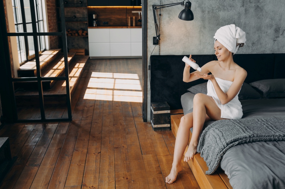 Morning skincare beauty ritual. Beautiful european woman wrapped in towel is sitting in bed. Slim girl applying body moisturizing lotion from bottle. Modern interior of bedroom. Bodycare and wellness.. Morning skincare beauty ritual. Beautiful european woman applying body moisturizing lotion.