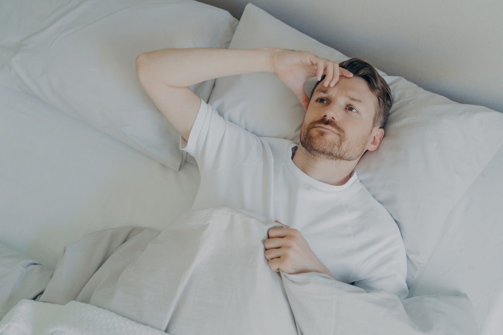 Young upset and tired bearded man lying on bed with open eyes and cannot sleep, male having insomnia problem or sleeping troubles. Bedtime and rest difficulty concept. Young tired bearded man lying on bed with open eyes