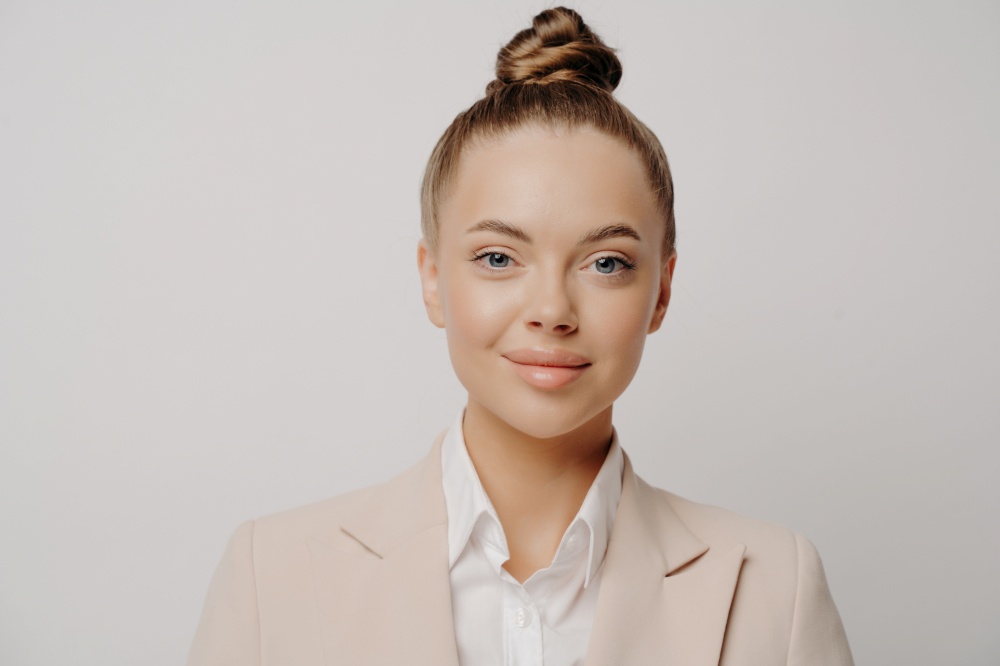 Close up portrait of young pretty brunette business lady with hair bun wearing formal clothes with white shirt standing isolated in front of grey background looking forward with confidence. Confident attractive business lady in beige suit