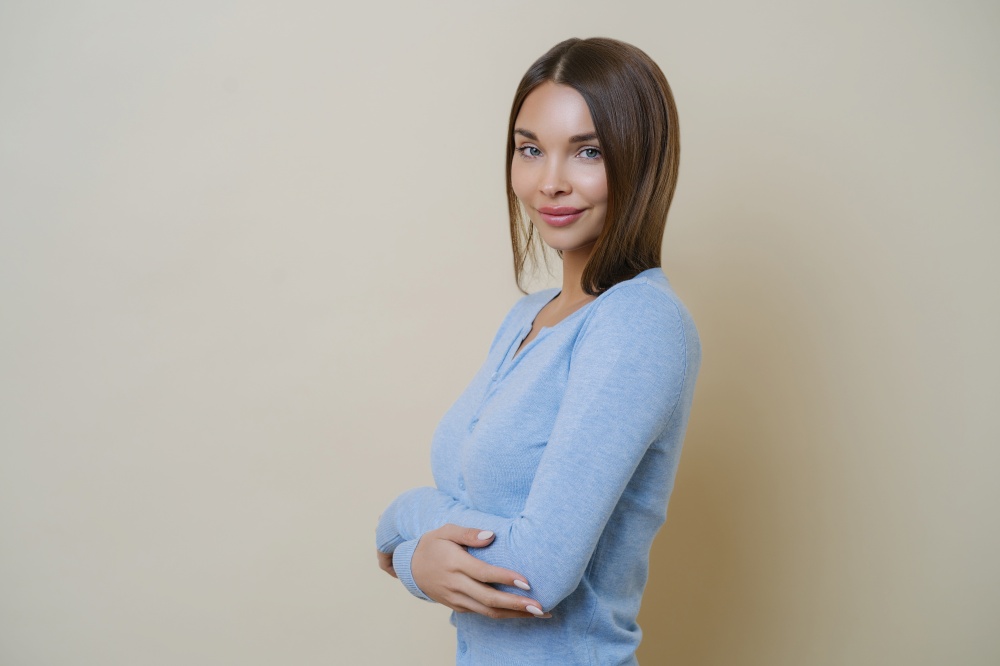 Charming slender woman with dark hair, keeps hands over body, has ideal appearance, smiles pleasantly, wears casual blue jumper, isolated over beige wall, returns from spa salon, had beauty procedures