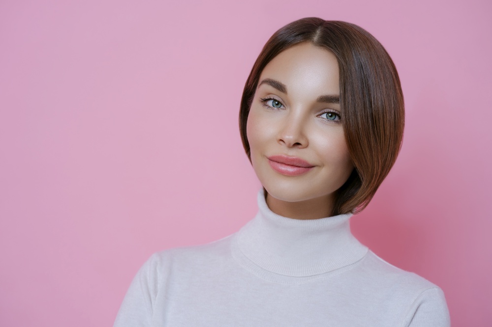 Portrait of good looking young woman with dark hair, minimal makeup, wears casual white turtleneck, going to have walk, poses against rosy studio wall, blank space for your advertising content.