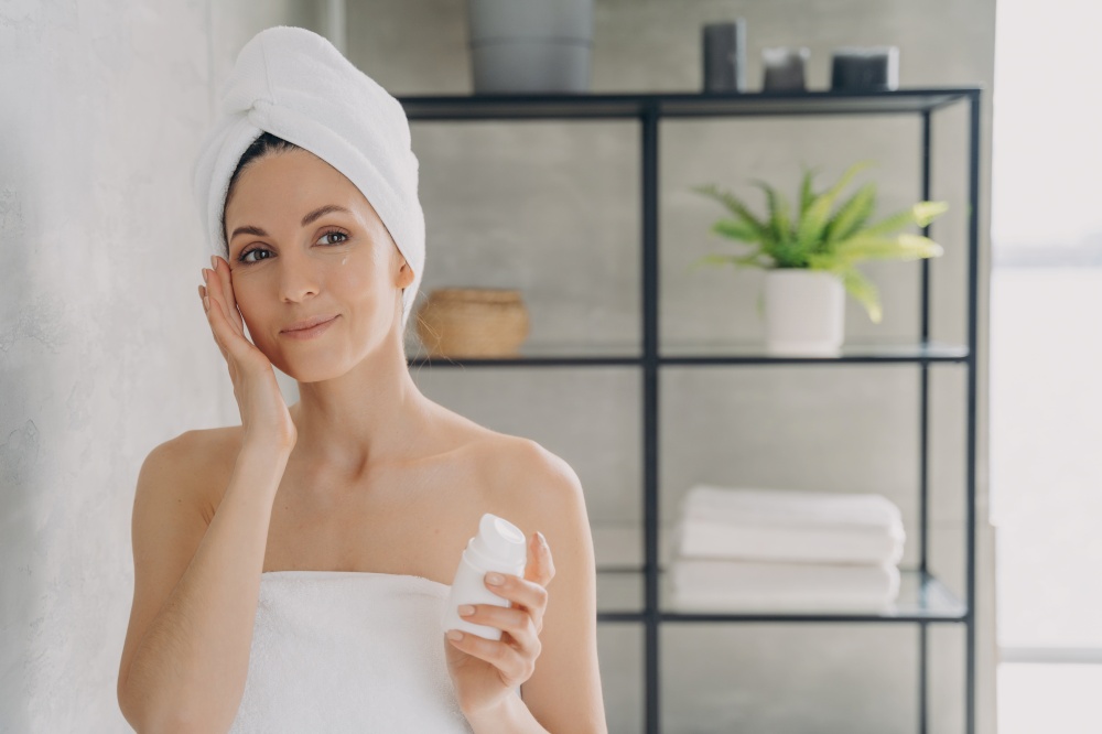 European lady applies face lotion or cream from bottle and looking into mirror. Face moisturizer, lifting cream. Attractive girl wrapped in towel after bathing and hair washing. Spa resort.. Face moisturizer, lifting cream. European lady applies face lotion or cream from bottle.