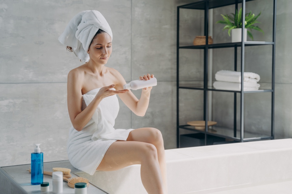 Attractive european woman wrapped in towel applying body lotion after bathing. Young woman takes shower in morning at home. Anti-cellulite massage and bodycare. Daily beauty routine and spa procedure.. Attractive european woman wrapped in towel applying body lotion after bathing. Daily beauty routine.