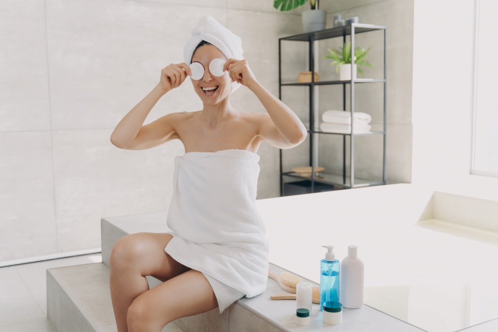 Happy young woman has fun applying face toner with cotton discs. Gorgeous european girl wrapped in towel after bathing. Lady is sitting on bathtub and relaxing. Woman takes shower at home.. Happy woman has fun applying face toner with cotton discs. Lady sitting on bathtub and relaxing.