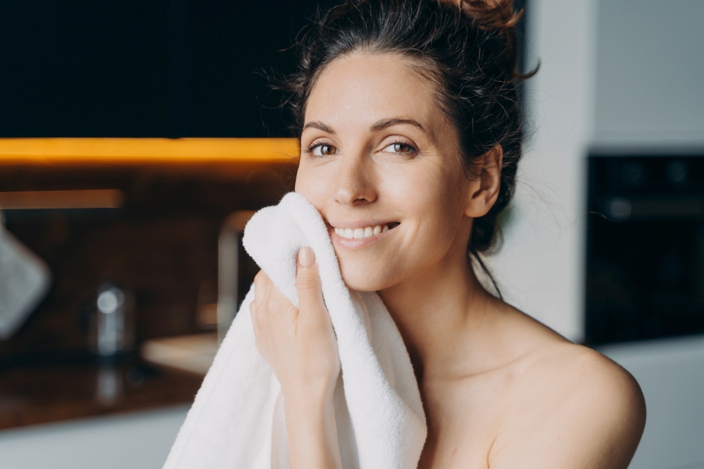 European girl is wiping face with towel after washing. Young brunette woman takes shower at home and doing skin care. Hygiene and freshness, dermatology and spa procedures.. European girl is wiping face with towel after washing. Young brunette woman takes shower at home.