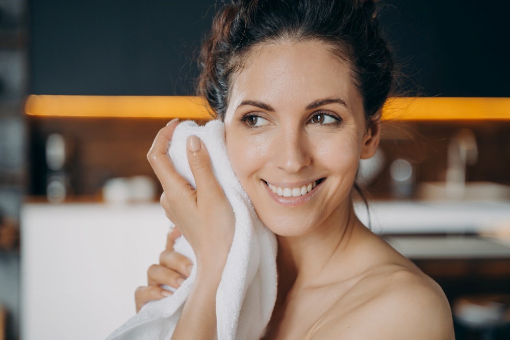 Evening beauty routine and hygiene. Caucasian woman is wiping face with towel after washing. Young happy woman takes shower at home. Face and body care, dermatology and spa procedures.. Evening beauty routine and hygiene. Caucasian happy woman is wiping face with towel after washing.