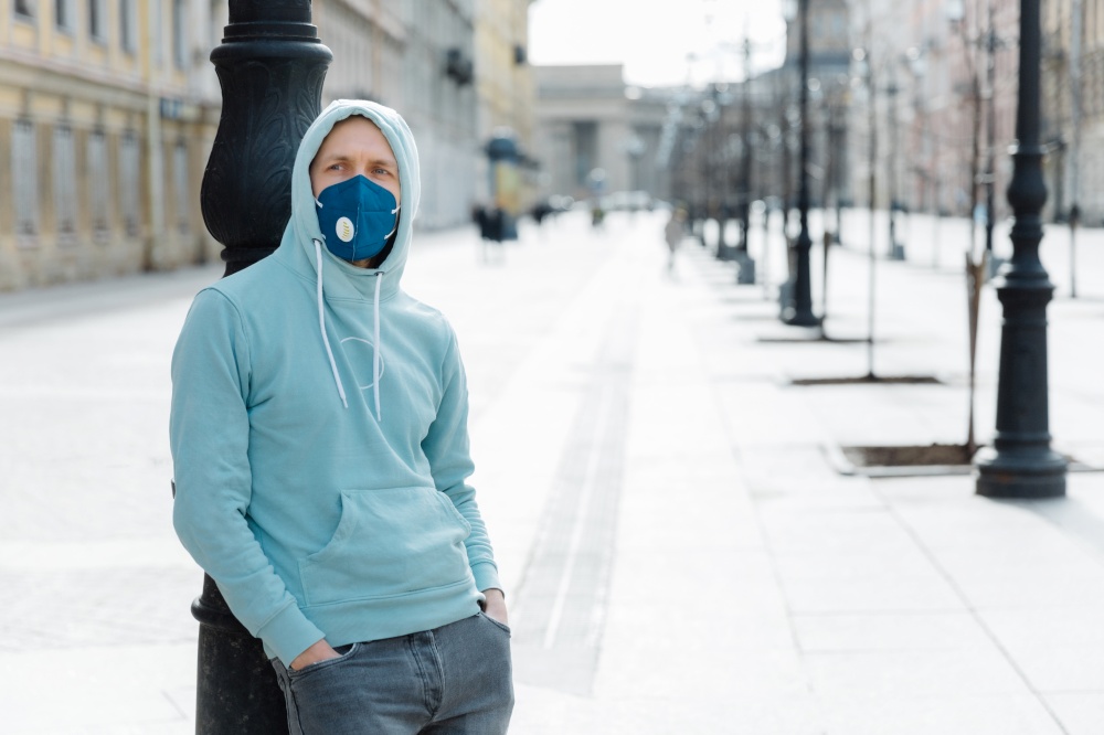 Pandemic coronavirus, Covid-19. Serious young man dressed in sweatshirt and jeans, wears respiratory mask to protect from virus or infection, walks through empty city streets during quarantine