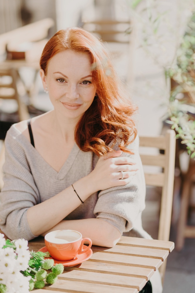 Vertical shot of attractive red haired young woman with wavy hair, dressed in warm jumper, drinks coffee or latte, sits at cafeteria outside, has pleasant conversation with boyfriend or lover.