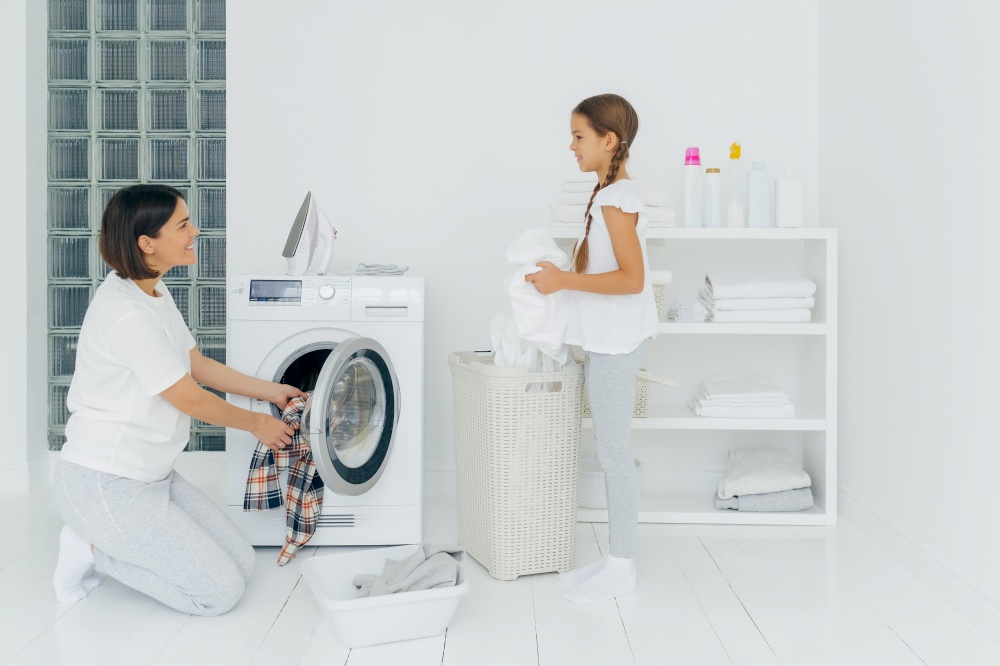 Glad mother and daughter busy doing laundry at home, have happy faces, brunette woman loads washing machine, little girl stands near basket with white linen. Housework, housekeeping concept.