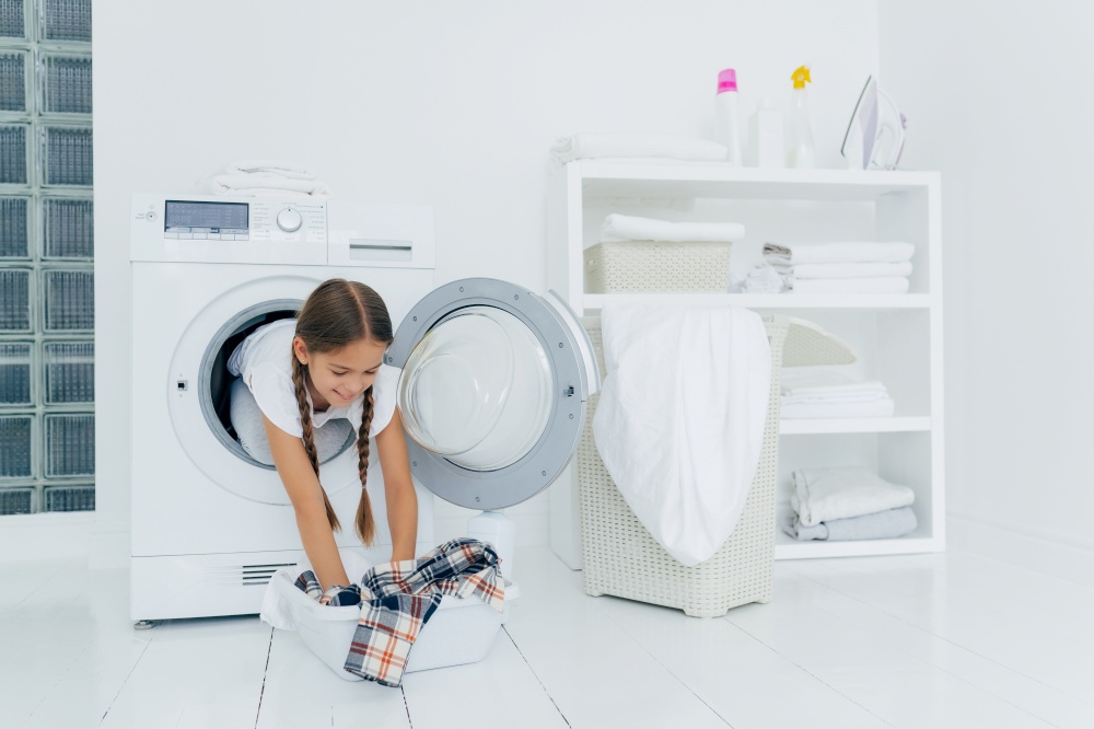 Pretty little girl householder poses inside of washing machine, takes checkered shirt from basin, engaged in laundry, has glad expression, two combed plaits. Childhood and washing day concept