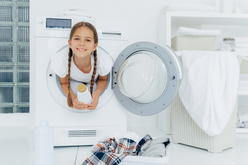 Smiling female child with glad expression, poses in washing machine, dressed in white t shirt, holds detergent, looks gladfully at camera, surrounded with much clothes and linen, does housework