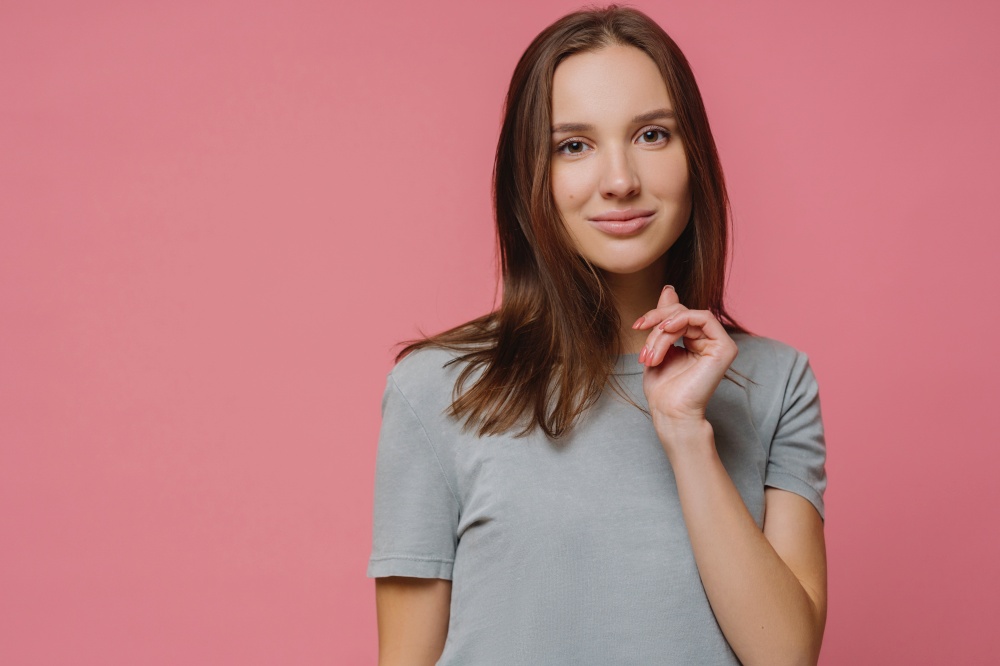 Confident lovely European woman has healthy pure skin, manicure, dressed in t shirt, enjoys photoshoot, models against rosy background with copy space, hears something pleasant or interesting