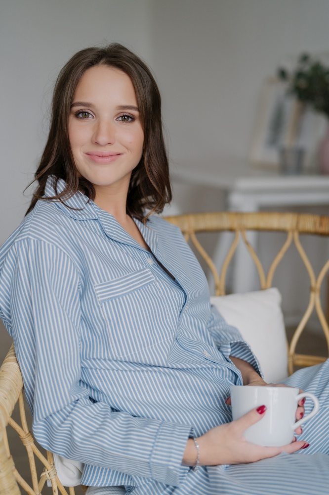 Satisfied beautiful woman with pleasant appearance, wears striped nightwear, drinks aromatic coffee, poses indoor. Relaxed housewife has drink at morning after awakening. People and rest concept