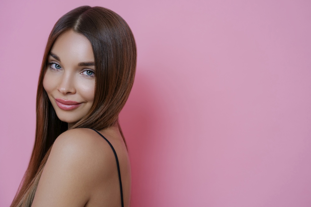 Portrait of beautiful woman with straight dark hair, clean skin, stands sideways shows bare shoulder, has pleased expression, models against rosy background, blank space on left for your advertisement
