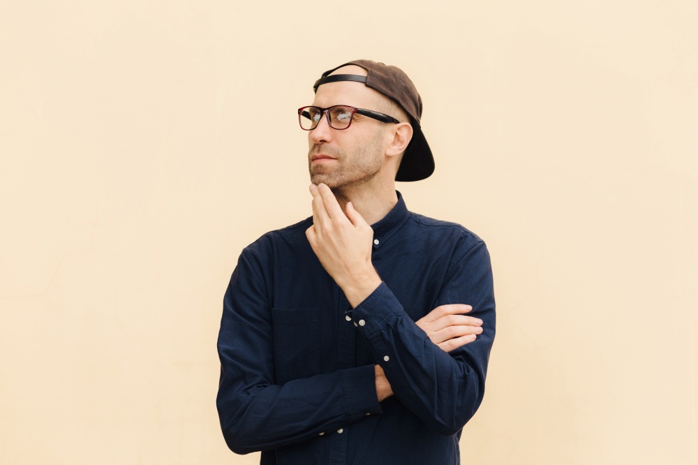 Indoor view of attractive middle aged fashionable male dressed in shirt and cap, looks pensively aside, thinks about something, poses against beige background. People, lifestyle, thoughts concept