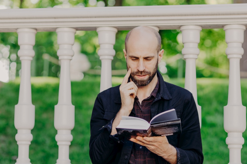 People, lifestyle, education, hobby concept. Attractive middle aged male with dark stubble, holds book, focused on page, reads informative information, poses outdoor, has serious expression.