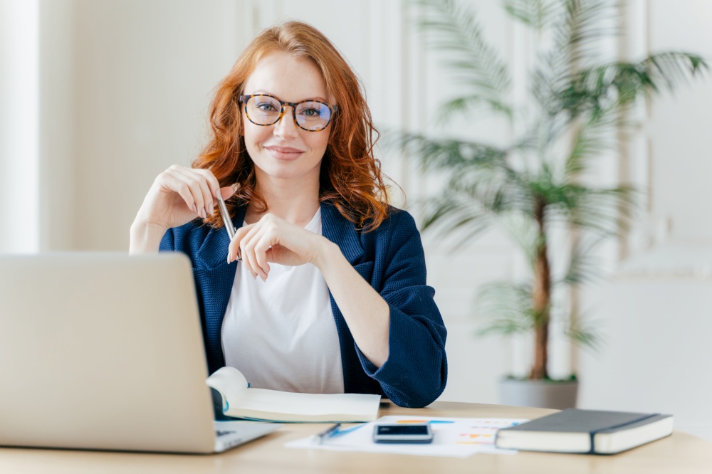 Positive red haired young female entrepreneur watches business webinar, writes email, makes notes in notepad, wears optical glasses, dressed in formal clothes, works online, involved in banking sphere