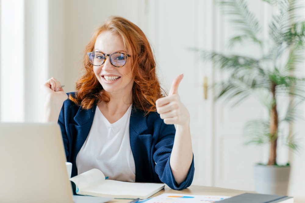 Positive redhead woman keeps thumb raised, demonstrates like gesture, satisfied with good work of colleague, updates software on modern gadget, searches data on website, poses in coworking space