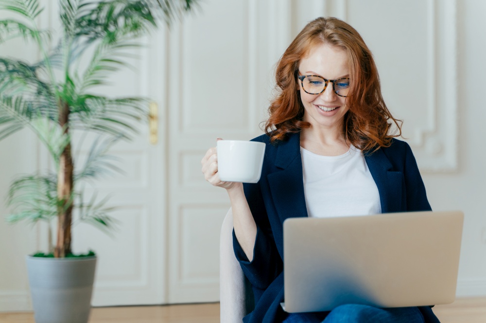 Satisfied European female with ginger hair, types information on laptop computer, has glad facial expression, drinks coffee, wears spectacles, develops new project, poses in cosy room alone.