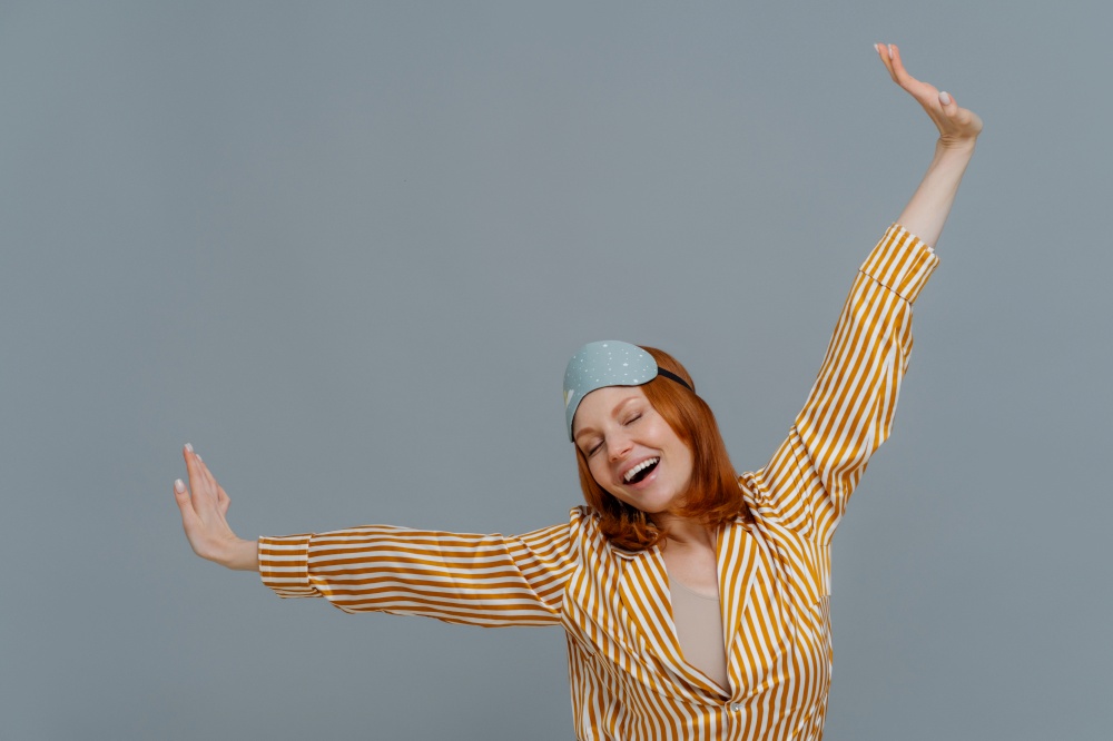 Good morning and awakening concept. Happy relaxed red haired young woman wears comfortable soft pajamas and sleepmask, stretches arms sideways, has glad expression, isolated on grey background.