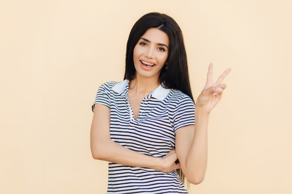 Portrait of cheerful brunette female with joyful expression, makes peace sign with two fingers, dressed casually, isolated over beige background. Beautiful woman shows victory gesture rejoices success
