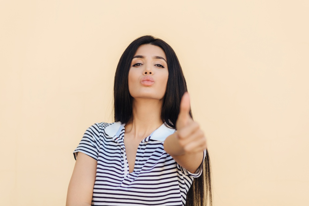 Confident pleased young woman with self assured expression, keeps thumb raised, shows approval gesture, has healthy skin, lips round, isolated over beige background. People and body language