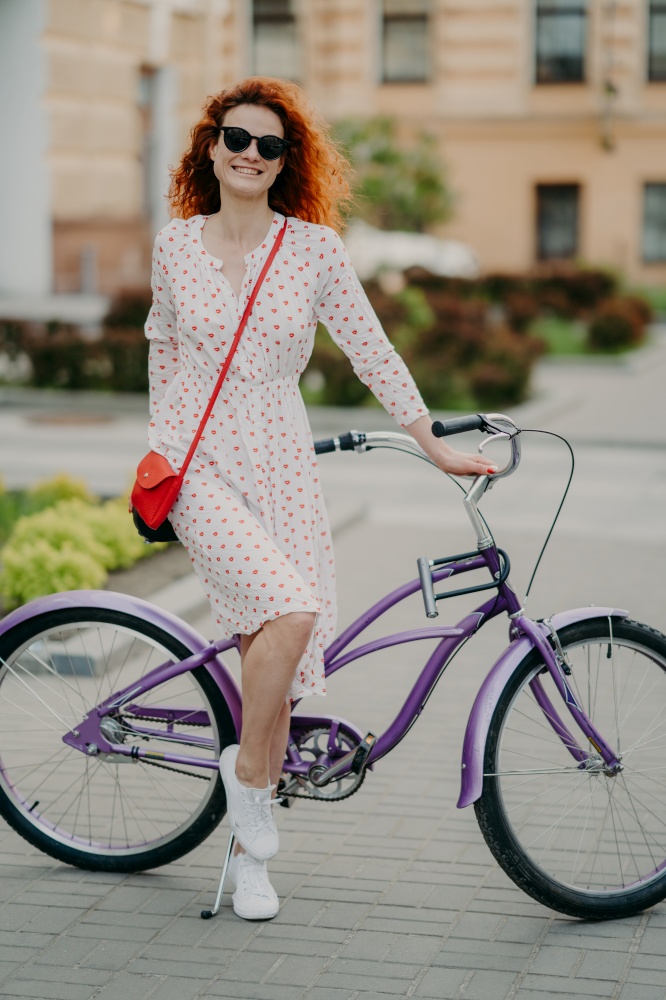 Pleased female has ride bike, dressed in fashionable outfit, smiles happily, stands near bicycle, being photographed in full length against blurred city background. Recreation and hobby concept