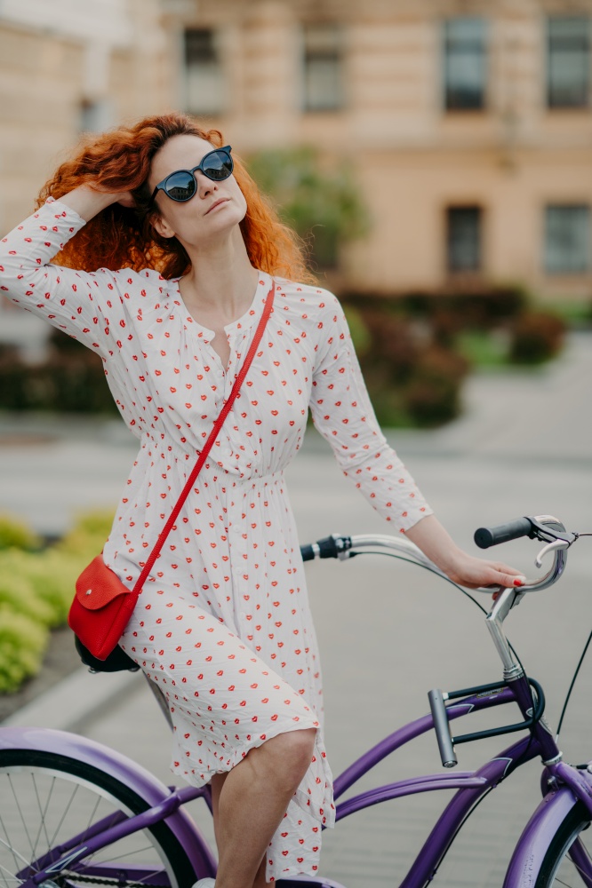Healthy outdoor activity concept. Lovely curly red haired woman with dreamy expression, uses bicycle for traveling across city, wears fashionable white dress and sunglasses, poses in urban setting