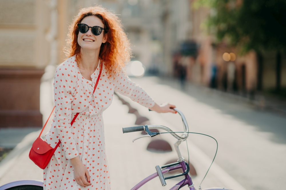Smiling red haired European woman has glad expression, stands near bicycle, carries red bag, focused somewhere into distance, poses over blurred background with copy space for your promotion