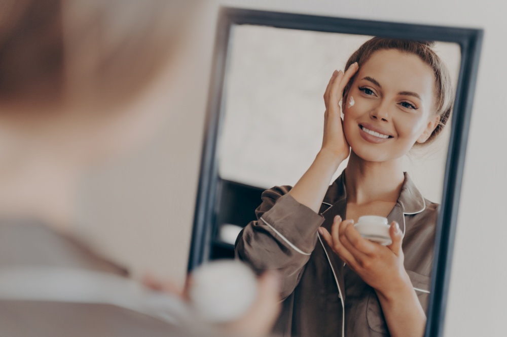 Positive happy young woman in pajama smiling, applying facial cream and looking in mirror, attractive lady doing morning beauty routine in bedroom at home. Skin care concept. Positive happy young woman smiling while applying facial cream